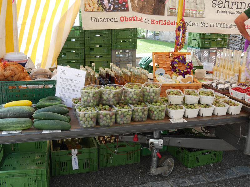 Ein Marktstand mit verschiedenen Obst- und Gemüsesorten, darunter Stachelbeeren, Gurken und Flaschen mit Saft, präsentiert Produkte in grünen Kisten und Körben.