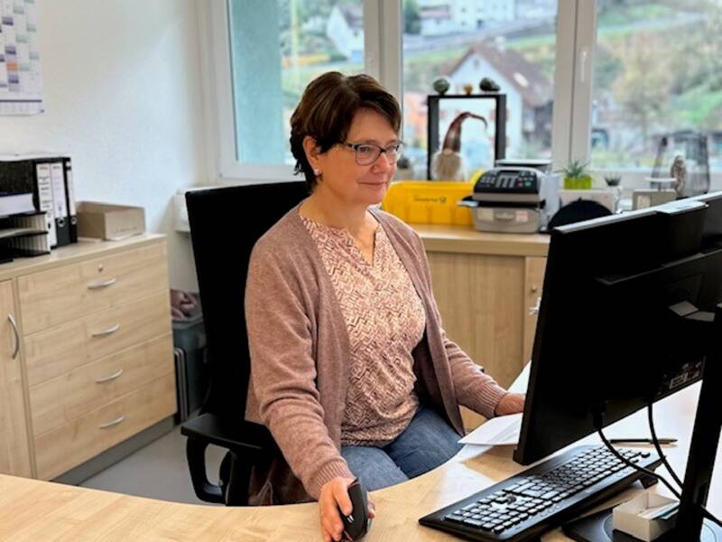 Eine Person sitzt an einem Schreibtisch und arbeitet am Computer in einem Büro mit Aktenordnern und einem Drucker im Hintergrund.